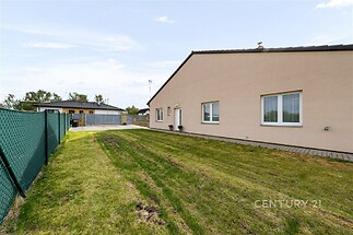 Zahrada s udržovaným trávníkem a pletivovým plotem vedle moderního rodinného domu s velkými okny, která poskytují dostatek denního světla