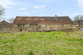 Historická kamenná stodola s červenou střešní krytinou, obklopena zeleným pozemkem. Nemovitost je vhodná k rekonstrukci v klidné venkovské oblasti.