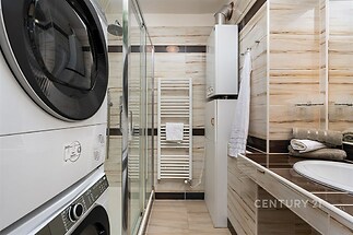 Koupelna s pračkou, světlé dlažby, moderní sprchový kout, úložný prostor, kombinovaný bojler, elegantní keramika, dobře osvětlená.