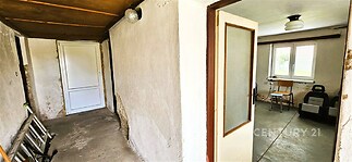 Chodba s betonovou podlahou a částí omítky, vedoucí do světlého pokoje s velkým oknem. Prostor je určen k rekonstrukci.