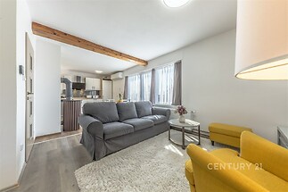 Světlý obývací pokoj s dřevěnými trámy, moderní kuchyní a parketovou podlahou, velkými okny a pohodlným sedacím nábytkem.