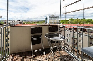 Malý balkon s dřevěnými dlaždicemi, výhledem na město a dvěma moderními židlemi nabízí příjemné posezení venku.