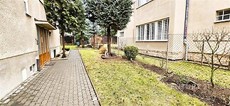 Úzká přístupová cesta s dlažbou, oplocená zahrada, stromy a keře, vedle bytového domu, vhodná pro moderní bydlení v klidné lokalitě.