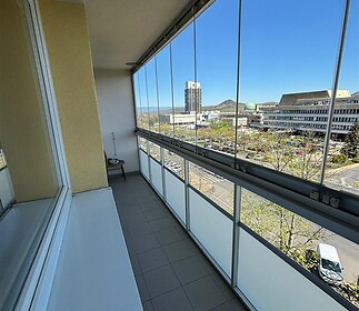 Uzavřená lodžie s velkými okny, dlažbou a výhledem na město, nabízí moderní prostor pro relaxaci.