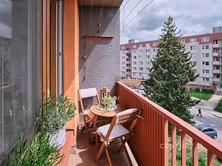 Balkon s dlažbou, výhledem do ulice a dvěma dřevěnými židlemi. Nachází se v panelovém domě a je ideální pro relaxaci nebo pěstování rostlin.