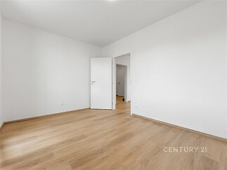 Světlý pokoj s parketovou podlahou a moderními bílými stěnami, vedle který je otevřený vstup do další místnosti.