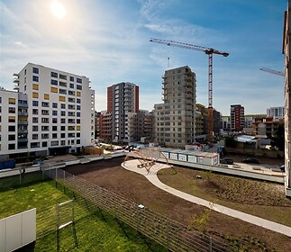 Výhled na moderní bytový komplex s balkony a parketovými chodníky, v blízkosti zelené plochy a dětského hřiště, ve výstavbě jeřáby v pozadí