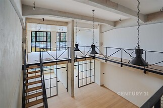 Moderní otevřený prostor s parketovou podlahou, velkými okny a industriálními prvky. Světlý interiér s kovovým zábradlím a schodištěm.