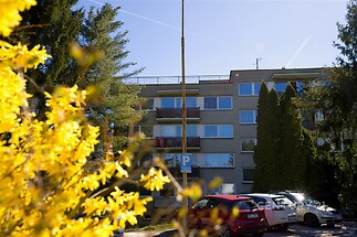 Exteriér bytového domu se světlými fasádami, velkými okny a balkony. Nachází se v klidné oblasti obklopené zelení a květinami.