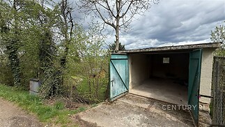 Prázdná garáž s betonovou podlahou a starými dřevěnými dveřmi, nachází se v klidném prostředí obklopeném zelení a stromy.