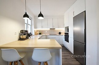 Moderní kuchyň s parketovou podlahou, světlým dřevěným pultem a vestavěnými skříněmi. Svítidla zavěšená nad barovým sezením.