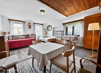 Světlý obývací pokoj s kamennou dlažbou, dřevěným stropem a velkými okny, obsahuje dřevěný nábytek a jídelní kout, působí útulně.