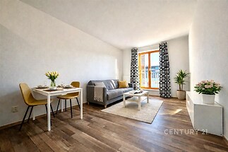 Světlý obývací pokoj s parketovou podlahou, velkými okny a moderním zařízením, poskytuje útulné prostředí k relaxaci a zábavě.