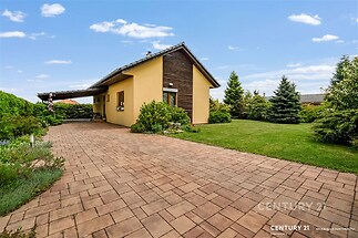 Předzahrádka s vydlážděnou cestou, udržovaným trávníkem a vzrostlými keři, moderní bungalov se žlutou fasádou a dřevěným obložením.