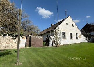 Tradiční venkovský dům s bílou fasádou, šikmou střechou a dřevěnými okny, obklopený zeleným trávníkem a kamenným plotem, ideální k rekonstrukci.
