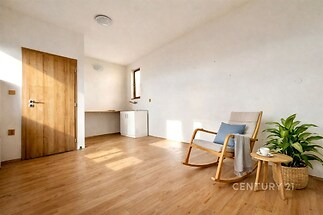 Světlý obývací pokoj s parketovou podlahou, moderním designem a velkým dřevěným křeslem, ideální pro relaxaci a odpočinek.