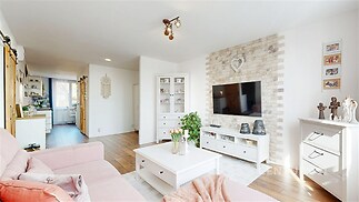 Světlý obývací pokoj s parketovou podlahou, moderní výzdobou a velkou televizí na zděné stěně, propojený s otevřenou kuchyní ve venkovském stylu.