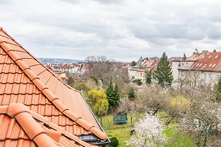 Výhled z podkrovního bytu na klidnou rezidenční čtvrť s červenými střechami, zahradami a stromovou zelení, ideální pro rodiny hledající příjemné prostředí.