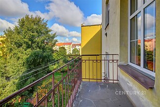 Balkón s kovovým zábradlím, výhledem na zelený park a historické budovy, světlý prostor s velkými okny, ideální pro relaxaci