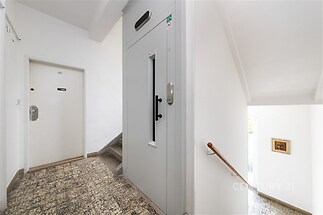 Chodba s výtahem a schodištěm, dlažba na podlaze, bílý nátěr stěn, čisté a udržované společné prostory budovy.