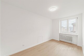 Světlý pokoj s dřevěnou podlahou, velkým oknem a bílými stěnami. Moderní interiér s radiátorem pod oknem.