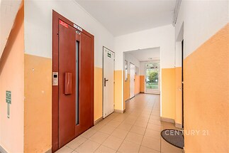 Vstupní hala bytového domu s dlažbou, červený výtah a světle oranžové stěny, blízko venkovních dveří.