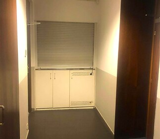 Úzká vstupní chodba s dlažbou, bílé stěny, vestavěné skříně a stropní osvětlení, moderní design, vhodné pro kanceláře.