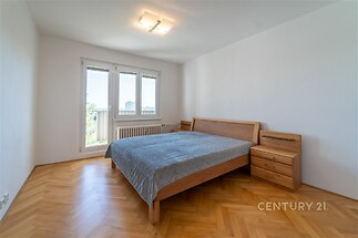 Prostorná ložnice s parketovou podlahou a velkými okny, moderní nábytek včetně dřevěné postele a nočních stolků, světlý interiér