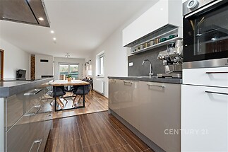 Moderní kuchyň s lesklými skříňkami, dřevěnou podlahou a vestavěnými spotřebiči, vedle ní prostorná jídelna s velkými okny.