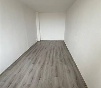 Úzký pokoj s moderní parketovou podlahou, čistě bílými stěnami a minimální výzdobou, ideální pro vestavěné skříně či osobní úpravy.