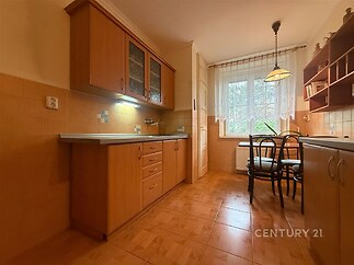 Kuchyň s parketovou podlahou a velkými okny, dřevěné kuchyňské skříňky a světlé obklady, jídelní kout s moderním osvětlením.