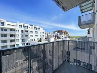Balkón s moderním zábradlím a výhledem na okolní bytové domy, dlažba a čistý design vytvářejí světlý a prostorný venkovní prostor.