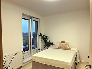 Světlá ložnice s velkými okny, které vedou na balkón, parketová podlaha a moderní čistý design který vytváří útulný prostor.