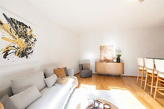 Světlý obývací pokoj s parketovou podlahou, moderním nábytkem a velkými okny, které zajišťují dostatek přirozeného světla.