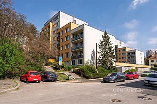 Fasáda bytového domu s moderními prvky, rekonstruovaná s novými balkonovými zábradlími, obklopena zelení a parkovacími místy.