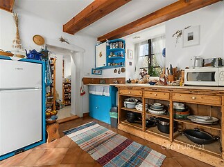 Tradiční kuchyň s dřevěnými trámy, dlažbou a vestavěnými skříněmi, nabízí útulný rustikální styl a světlé prostředí díky velkému oknu.