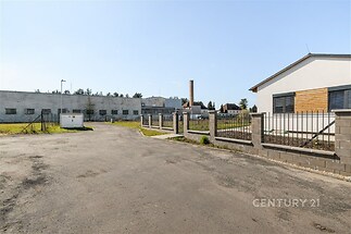 Příjezdová cesta k modernímu průmyslovému objektu s dlážděným povrchem a oplocením, v klidné lokalitě blízko zeleně