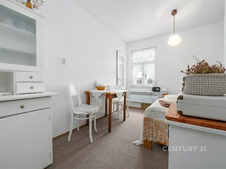 Jídelní kout s bílým nábytkem, kobercovou podlahou a velkými okny poskytujícími dostatek světla, vytváří světlý a útulný prostor.