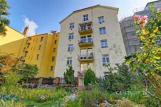 Vícepatrový bytový dům s balkony, zahrada v přední části. Budova má žlutou fasádu a nachází se ve městě.