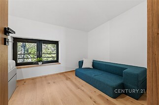 Minimalistický pokoj s parketovou podlahou, moderní designový gauč a velká okna s výhledem do zeleně, ideální pro odpočinek