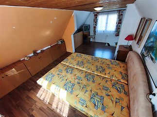 Podkrovní ložnice s dřevěným stropem, parketovou podlahou a vestavěnými skříněmi, nabízí útulný prostor s velkými okny pro přirozené světlo.