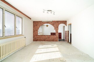 Světlý obývací pokoj s velkými okny a cihlovou stěnou, dřevěná podlaha chybí, vhodný pro rekonstrukci, poskytuje potenciál pro moderní prostor.