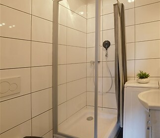 Malá koupelna s moderním sprchovým koutem, dlažba a místo na pračku. Světlý prostor s jednoduchým bílým obkladem.