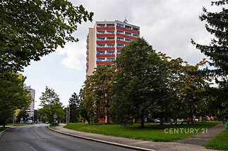Vysoký bytový dům s balkony a barevnou fasádou, obklopený zelení a chodníkem v klidném městském prostředí.