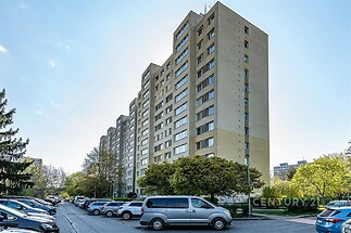 Vysoký obytný panelový dům, renovovaná fasáda, parkoviště před budovou, vzrostlé stromy, klidná lokalita, výhled na město.