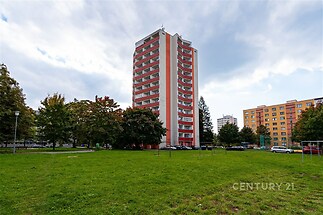 Vysoký bytový dům s balkony, obklopený zeleným parkem a stromy, poblíž parkování. Budova je moderní a nachází se v klidné lokalitě.