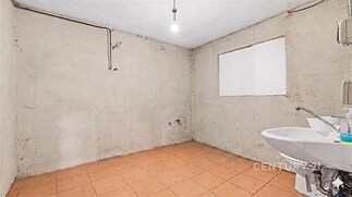 Prázdná místnost se světlou dlažbou a umyvadlem, vhodná pro rekonstrukci, s neomítnutými stěnami a základním osvětlením.