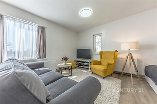 Světlý obývací pokoj s parketovou podlahou, moderní šedou pohovkou a velkými okny, které zajišťují dostatek přirozeného světla.