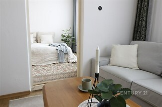 Světlý obývací pokoj s parketovou podlahou, moderní pohovkou a výhledem do útulné ložnice s velkým oknem a světlým dekorem.