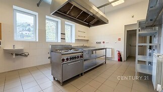 Profesionální kuchyň s dlažbou, nerezovými spotřebiči a velkými okny. Světlý prostor s moderními prvky a účelným designem.